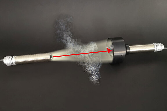 Zwei industrielle zylindrische Sensoren, verbunden durch einen roten Pfeil, der einen Lichtstrahl durch eine Rauchwolke anzeigt.